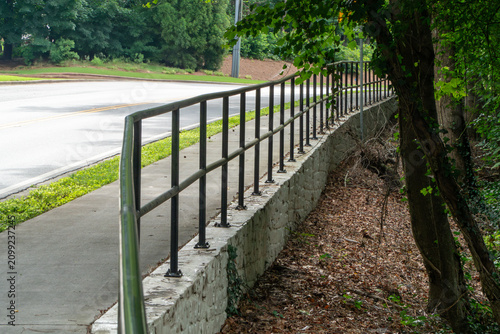 Картината върху платно A curved concrete sidewalk with a sturdy black metal railing overlooking a tranquil forest floor of brown leaves, bordered by an asphalt road and lush green trees on a bright day