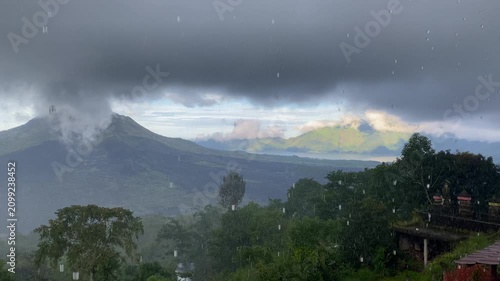 Wide view of lush green hills and misty mountains at Kintamani volcano in Bali, Indonesia during heavy rainfall, with a lake visible in the background.