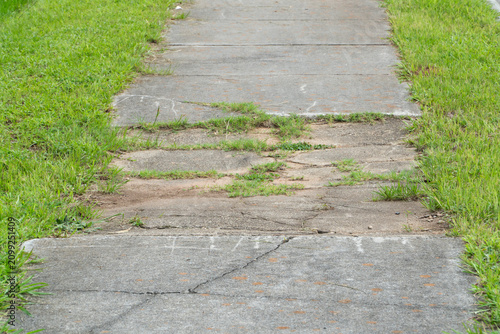 Overgrown and heavily cracked gray concrete footpath, flanked by vibrant green grass, demonstrating wear, tear, and natural encroachment in an outdoor setting.