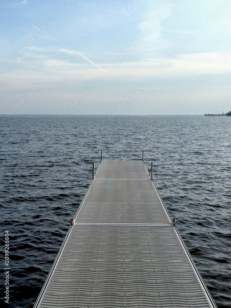 Fototapeta premium Long floating dock stretching into calm open water beneath a soft blue sky with wispy clouds.