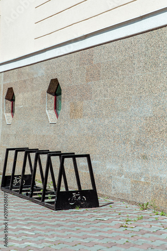 Black metal delta-style bike rack with five slots and bicycle iconography against a speckled granite wall, urban cycling infrastructure on patterned brick pavement, public bike parking solution.