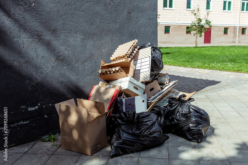 Pile of discarded cardboard boxes and black plastic trash bags against a dark textured wall near a residential building, urban waste accumulation, post-delivery debris in a city courtyard