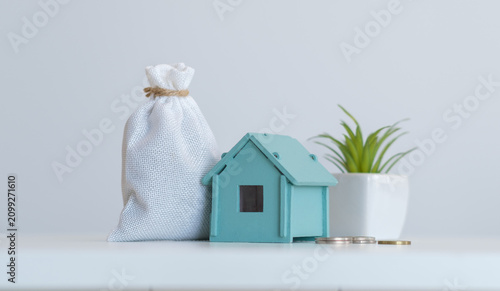 Conceptual financial image featuring a miniature blue house model, a white money bag, and coins, representing savings for real estate, home ownership, or mortgage planning.