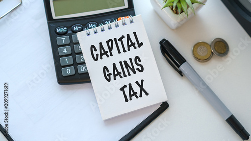 Conceptual financial photography featuring a notepad with the words CAPITAL GAINS TAX written on it, placed on a white desk next to a calculator, a pen