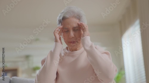 Elderly senior woman touching her neck and shoulder in severe pain while sitting on a sofa at home. Concept for neck ache, muscle strain, cervical spine issue, and senior healthcare.