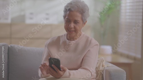 Older asian woman using mobile phone sitting relaxed on couch at home, smile or laugh with happy time. enjoy watch or read text on social media app. Senior asian with digital technology after retired.
