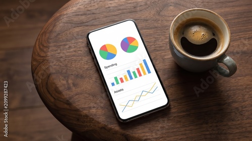 Smartphone on wooden table displaying colorful financial charts and graphs next to coffee cup, mobile finance app and casual workspace setup for analytics