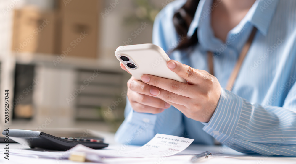 Fototapeta premium Close-up of female employee hand using smartphone with financial calculator and invoice receipt in office. Professional business woman checking mobile payment and online banking data.