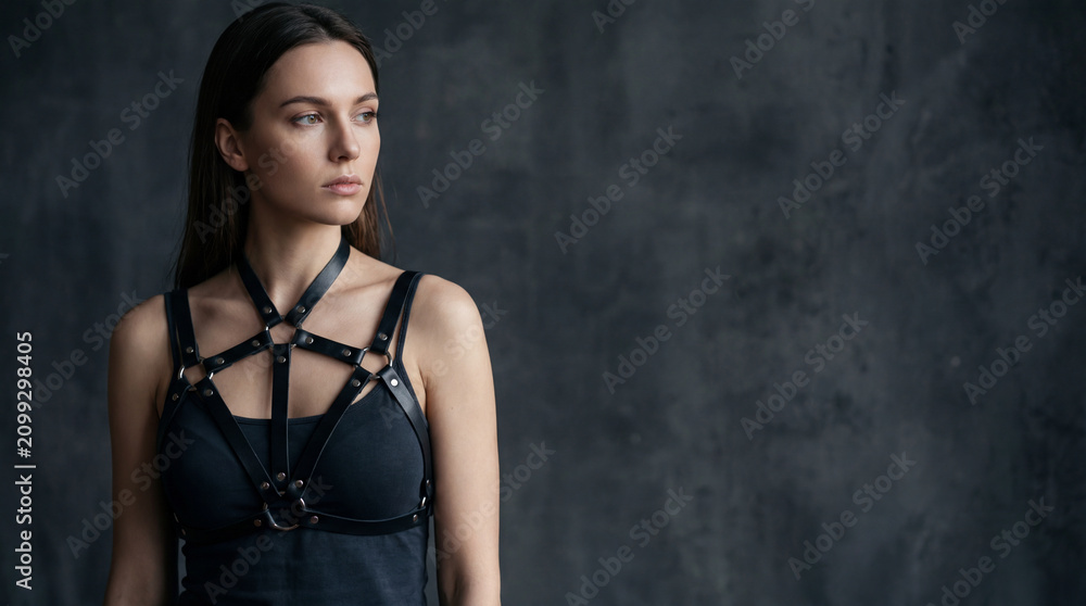 Fototapeta premium young brunette woman wearing black top with leather harness near dark textured wall looking aside with confident calm mood for alternative fashion portrait with copy space for style branding