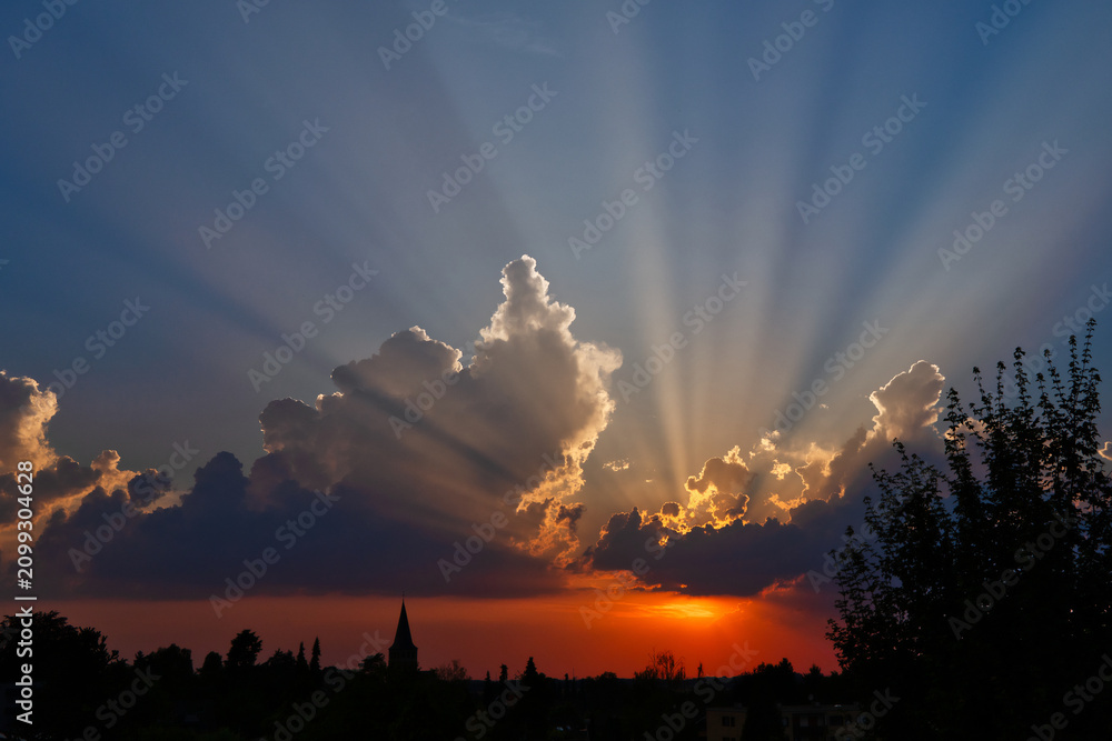 Fototapeta premium sunset with the silhouette of a church and dramatic, colorful cumulus clouds with sunbeams over Ratingen Homberg