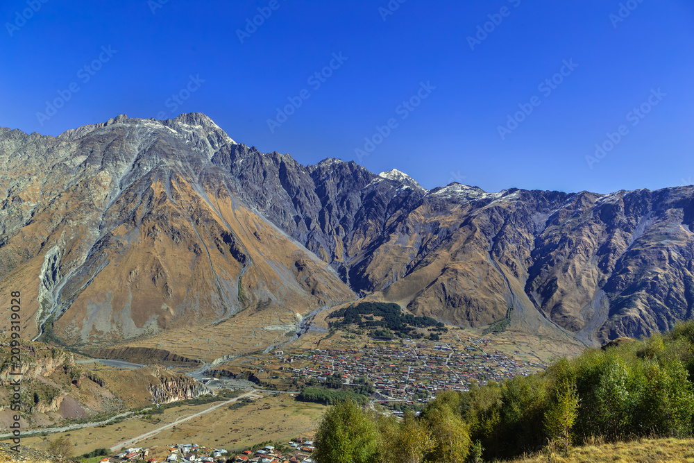 Fototapeta premium High angle view of Stepantsminda town in the Caucasus mountains, Georgia. Scenic panorama from a hill in sunny daylight