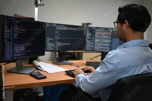 Focused Asian male software developer types on a keyboard analyzing complex programming code on multiple computer monitors while working at a desk inside a modern corporate IT business office.