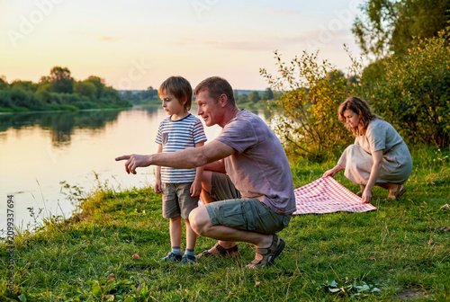 Man shows river view to little boy while woman prepares picnic at sunset.