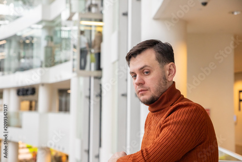 Thoughtful man in orange sweater looking away in shopping mall. Pensive male person leaning on railing in indoor urban space. Concept of lifestyle, waiting and contemplation in public area.