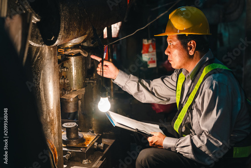 Engineer helmet safety vest inspection machine factory worker clipboard industrial maintenance male technician examining heavy equipment with lamp during night shift in dim manufacturing workshop