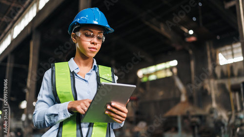Engineer worker tablet helmet vest factory industry woman safety inspection, confident female industrial worker using digital tablet in factory while wearing blue hard hat and green safety vest
