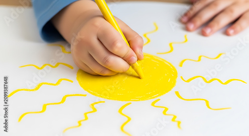Child holding yellow crayon drawing a sun on white paper
