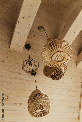Low angle view of modern eco-friendly pendant lamps made of rattan, jute and metal rope hanging from a light wooden plank ceiling in a cozy, stylish interior