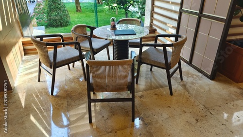 A table and wicker chairs are placed on the tiled floor near the large window. Nearby, there are trees and a flower bed on a grassy lawn