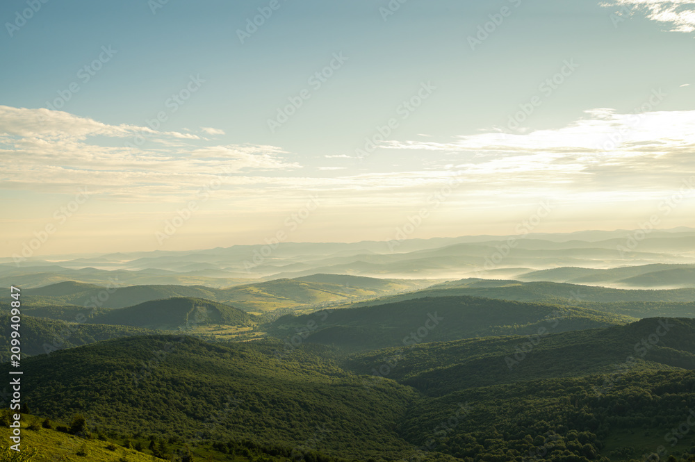Fototapeta premium Scenic misty mountains with wind turbines at sunrise. Renewable energy farm in foggy green hills. Beautiful landscape of sustainable power and nature conservation