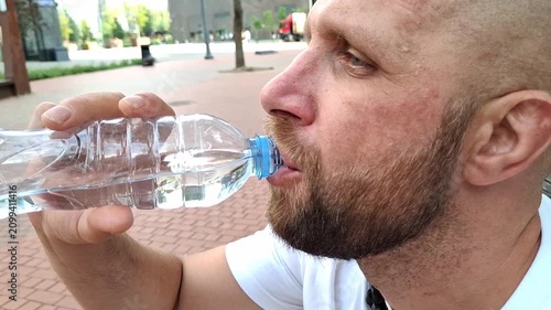 Side view of man holding plastic water bottle outdoors on city street, hydration and urban lifestyle in summer. Guy drinks water on a hot day