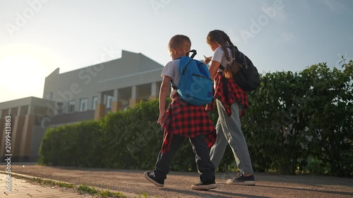 Student siblings walk along street in sunlight. Girl and boy walking hand in hand near school building. Morning road scene with backpack child walk. Student walk to school holding sibling hand