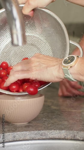 Vertical video: Tilting colander under faucet, hands draining tomatoes into pink bowl with watch