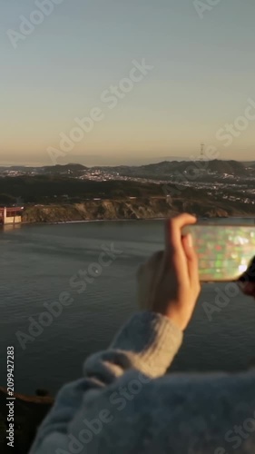 Vertical video: Seeing sun, woman in gray knit framing Golden Gate on phone, overlook, copy space