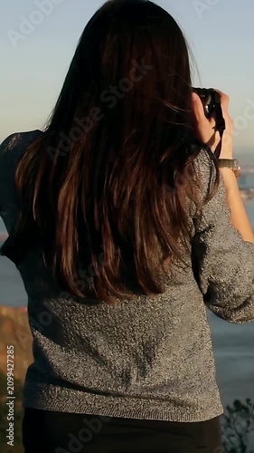 Vertical video: Sun drawing woman grey sweater raising camera to eye on cliff, shooting sea view