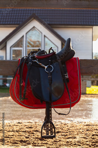 horse saddle hangs on wooden fence with house in background