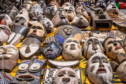 A shot of a large variety of traditional ethnic masks, mostly handcrafted from wood with African tribal designs, displayed for sale on a yellow patterned cloth at an outdoor market stall.