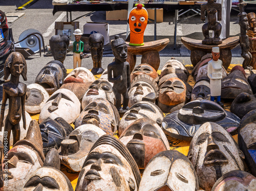 A shot of a large variety of traditional ethnic masks and idols, mostly handcrafted from wood with African tribal designs, displayed for sale on a yellow patterned cloth at an outdoor market stall.