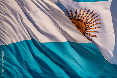 Close-up of the Argentine national flag waving in the wind under a clear blue sky. The image highlights the blue and white stripes and the Sun of May, symbolizing Argentina's national identity.