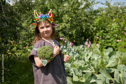 A primary school-aged girl holds forks of broccoli against a backdrop of a vegetable garden with cabbage beds. She wears a homemade headband made of fuzzy wire with goat ears and horns.