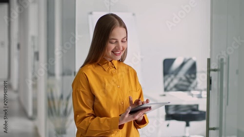 Smiling Young Caucasian Woman Engaged While Using Tablet In Modern Office. Happy Female Professional Holding Device, Working In Bright And Stylish Workspace, Excited About Task At Hand.