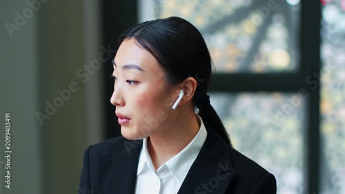 Focused Asian Woman Wearing Wireless Earbuds Engaged In Conversation While Sitting At A Modern Office. Professional Environment With Clear Focus On Work And Productivity, Business Concept.