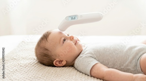 Digital infrared thermometer measuring temperature of a baby. Infant lying on a blanket during a health check. Pediatric healthcare and fever monitoring concept