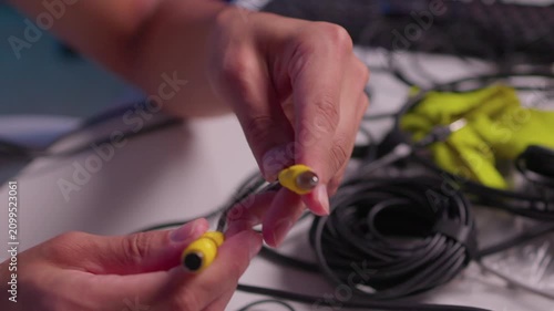 Hands aligning yellow electrical connector halves on black cable over tabletop workspace, showing careful joining motion, exposed pin, coiled wire, green fabric backdrop