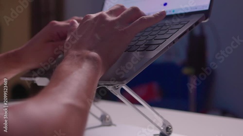 Hands typing on laptop raised by foldable metal stand on white desk, showing ergonomic workstation setup, active keyboard use, slight forward tilt, compact modern office scene