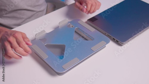 Hands unfolding and positioning metal laptop stand on neutral desk for ergonomic workspace setup beside closed laptop and tablet, showing careful adjustment, placement, and practical desktop