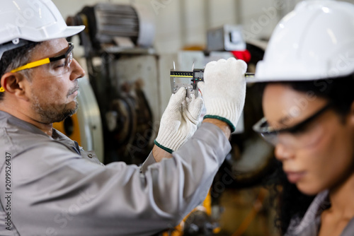 Industrial workers in hard hats measure a part with calipers, wearing safety gear on a busy factory floor, focused on precision and quality control.