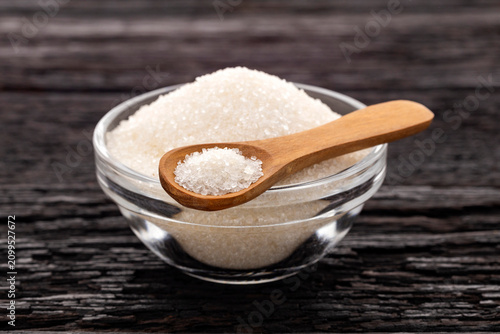 Close-up of refined sugar crystals in a clear bowl with a wooden spoon. Stock photo related to baking, sweeteners, gastronomy, and food preparation.