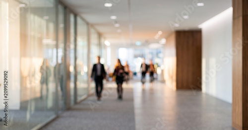 A modern office corridor with blurred figures walking. The space features glass walls and wooden accents, creating a professional ambiance.