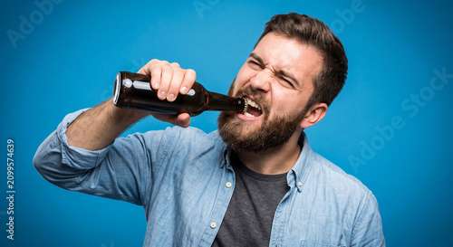 Humorous Bearded Man Trying to Open Glass Beer Bottle With Teeth Over Blue Background