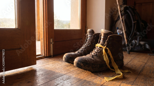 Pair of muddy hiking boots resting on a wooden floor near an open door with a backpack and walking stick in the background