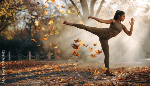 A woman practicing yoga outdoors during autumn, balancing on one leg with falling leaves around her in a park-like setting with sunlight filtering through trees.