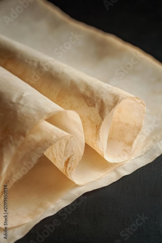 Rolled parchment paper with light brown staining and creased edges is presented on a dark surface. AI.