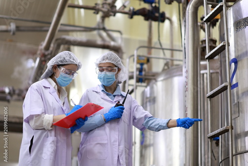 Female engineers inspecting industrial storage tank and piping system in food factory, Quality control specialists auditing production standards near large stainless steel vats in industrial plant. 