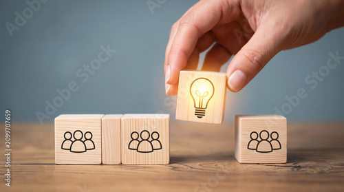Hand inserting a wooden block with a glowing lightbulb among team icons, representing business innovation, creative solutions, and leadership