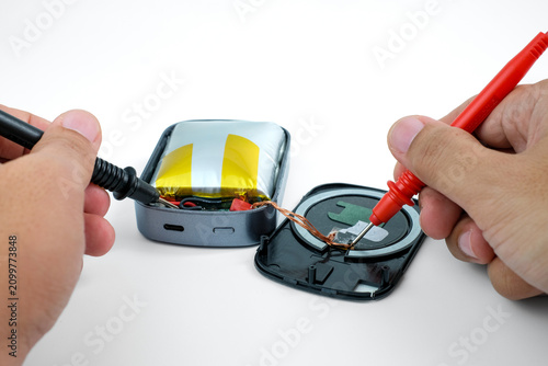 Hands use multimeter probes to test an opened power bank with a swollen lithium battery and exposed wiring on a white background. Suitable for testing damaged power bank, battery diagnostics